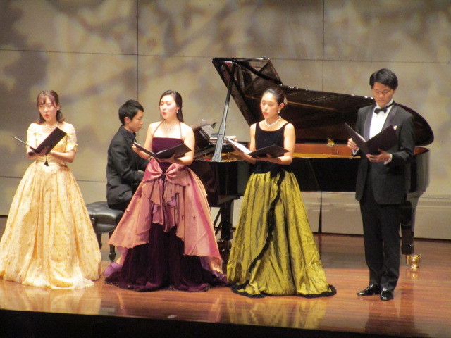 舞台上でピアノ奏者と歌手4人が音楽を披露中。
