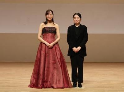 舞台上に二人の女性が立っている写真。片方は赤と黒のドレス、片方は黒のパンツスーツ。
