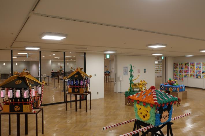 展示スペースにディスプレイされた、児童が飾り付けをおこなったカラフルなミニ御おみこし3台とその奥に見える別の展示物と絵画の画像。