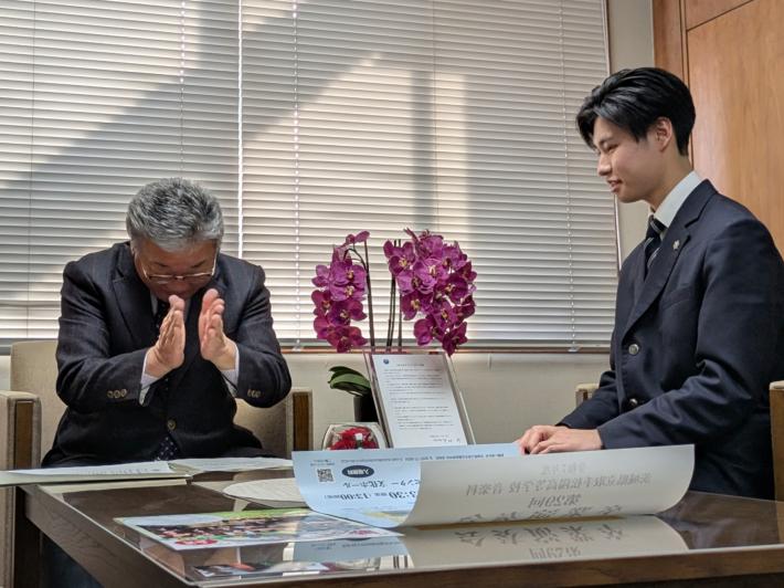 椅子に掛けて対談している2人の男性。一方は制服姿の高校生、もう一方はスーツ姿の成人男性で拍手をしている