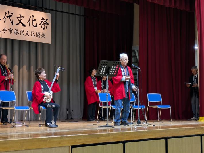 赤い法被を着た男女がステージ上で、三味線、歌、尺八で演奏している様子
