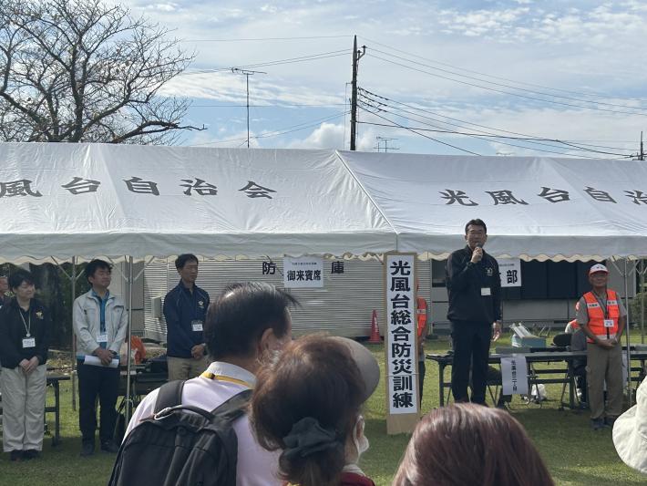 光風台総合防災訓練と書かれた看板の横からあいさつをする男性
