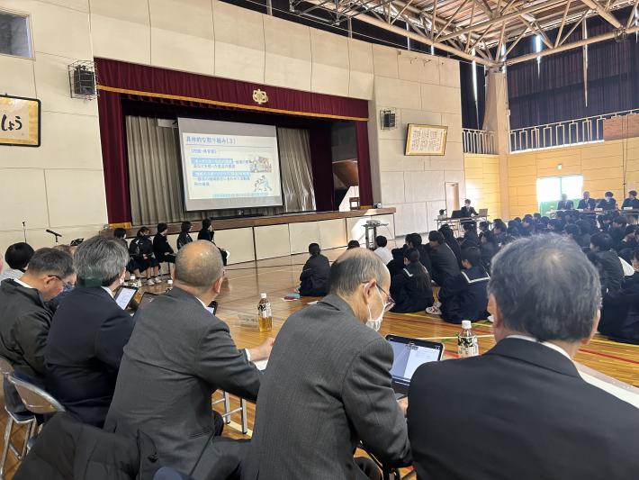 体育館前方に設置されたスクリーンに映った動画をみる生徒たち