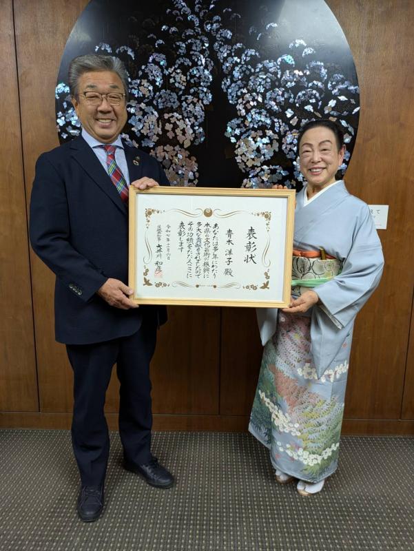 スーツを着た男性と着物を着た女性が表彰状を中心にもって立っている。