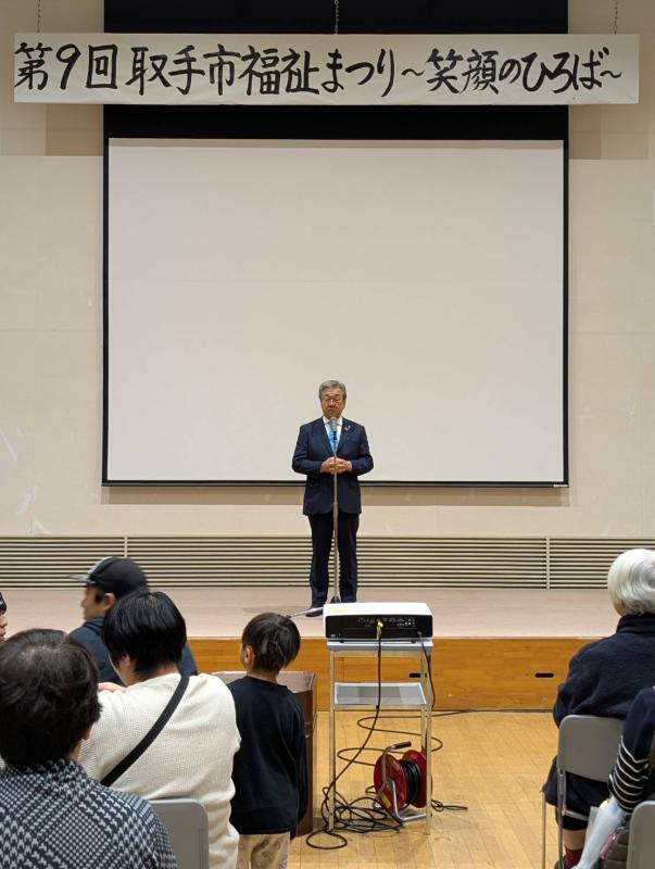 取手市福祉まつりと書かれた看板の下からあいさつをする男性