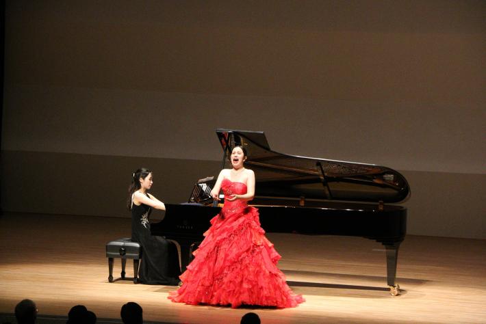 ドレスを着て歌う女性と、ピアノを弾く女性