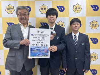 賞状を手に持つ大人の男性と男子学生。その横に男子学生が立っている。