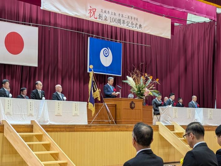 取手第二高等学校100周年記念式典と書かれた看板の下から挨拶をする男性