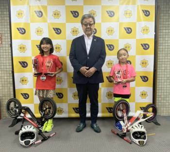 ランバイクを下に置き、パネルの前に立つ小学生2名とその中央に立つ男性