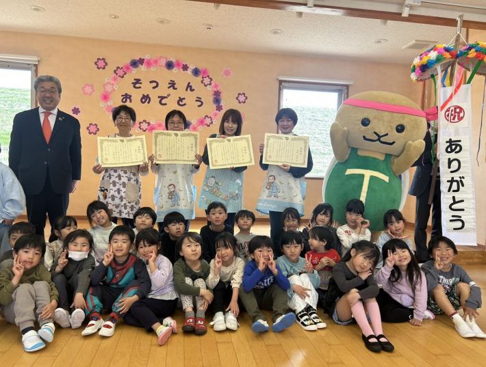 立って感謝状を持つ複数の保育士と、床に座るこどもたち