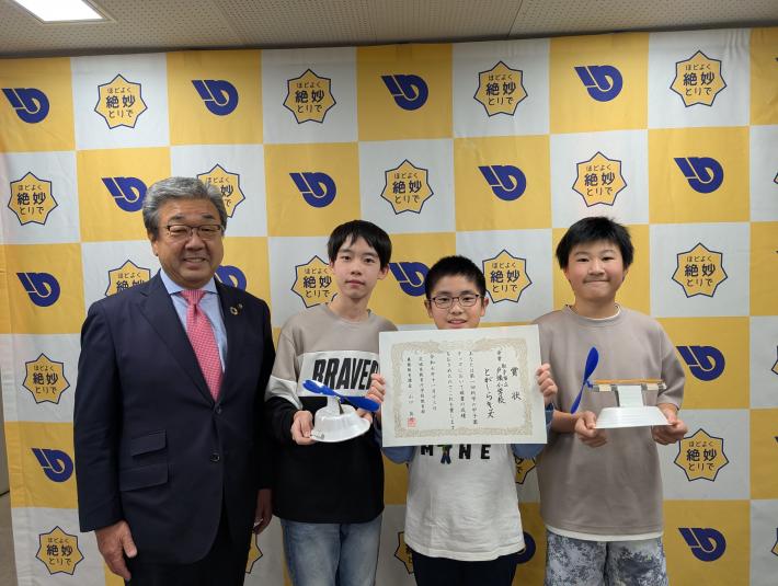 賞状と作品を持つ小学生3人と、その横に立つ男性