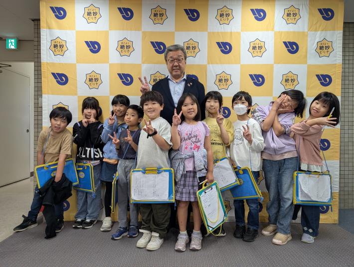 パネルの前でピースをする男性と小学生たち