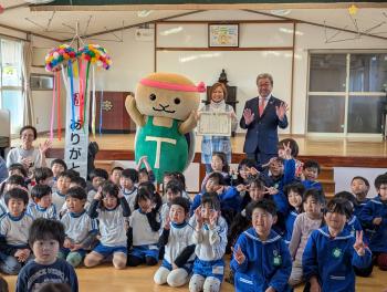 こどもたちに囲まれた保育士が表彰状を持って立っている。その隣には、スーツを着た男性がいる。