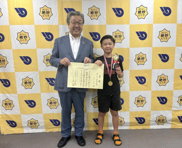 パネルの前で賞状とトロフィーを持つ小学生と男性