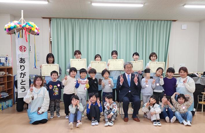 床に座るこどもたちと、表彰状を持って立っている複数の保育士