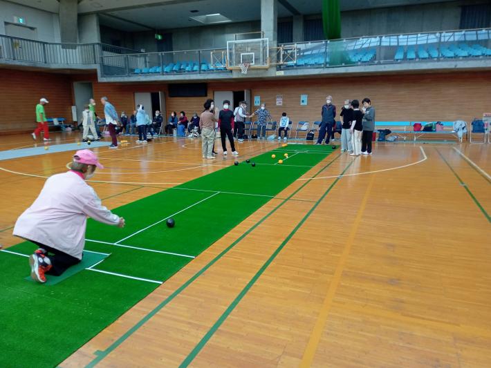 体育館内において、参加者が床に敷かれたシートの上でボールを転がしている様子