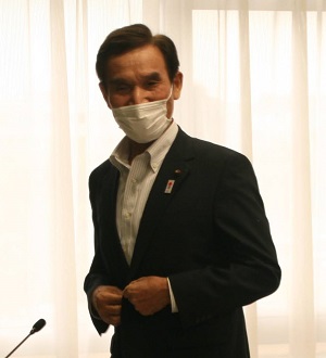 黒いスーツを着て白いマスクをつけた短髪の男性がスーツのボタンをとめながら立ち、あいさつをしている