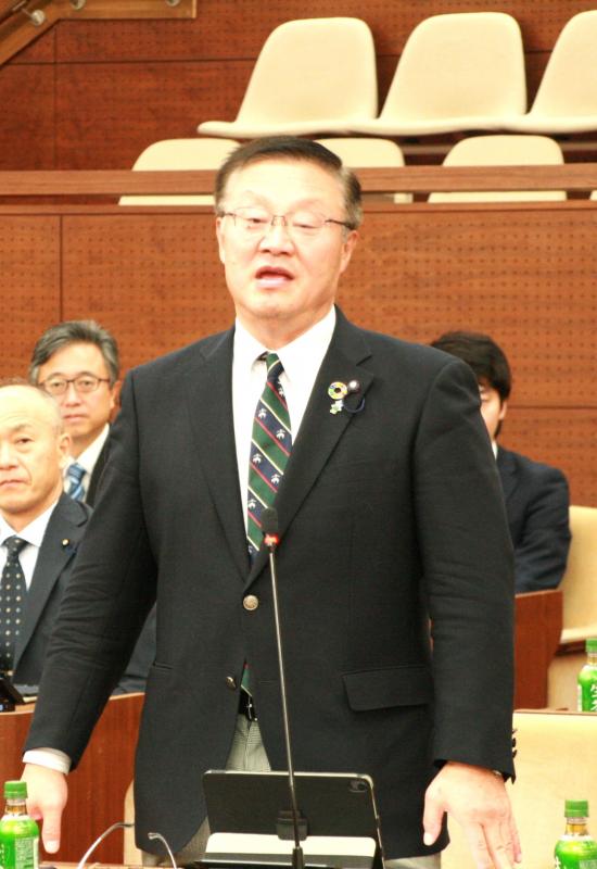 議場にて紺色のスーツを着た男性が起立してあいさつをしている