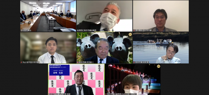 8分割にされたZoom画面に、それぞれ会議室と、スーツを着た男性7人が映っている。会議室には14人の男性と女性が座っている