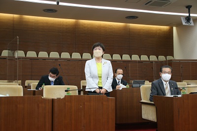 白いスーツを着た女性が議席脇に立ち挨拶をしている。周りには間隔を開けて3人のスーツを着た男性議員が着席している