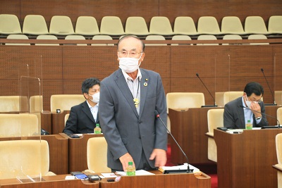 議席にて、マスクを着用しスーツを着た男性がその場で起立し、挨拶をしている写真