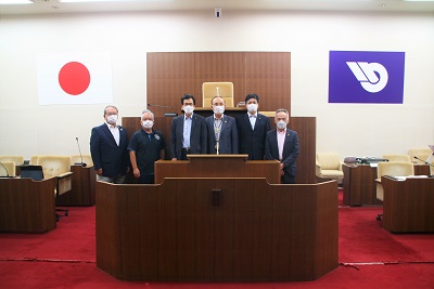 壇上にて、スーツを着た男性6人が横一列に並んでいる。左から赤羽議員、金澤議長、延岡市議会議員4人。