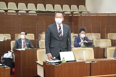 議場の議席にて起立しあいさつをするストライプ柄のネクタイを身につけた眼鏡の男性委員長。後ろには2人の男性が映る