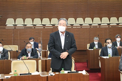 議場の議席最前列中央で立ち、あいさつする眼鏡をかけてスーツを着た男性議員。後ろには男性が6人映る。
