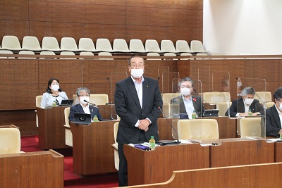 議席最前列で起立しあいさつする眼鏡をかけた男性委員長。周囲には間隔を開けて座る議員と職員が5人映る