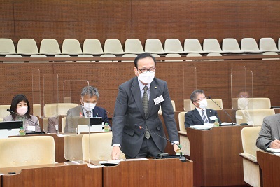 議席最前列中央で起立しあいさつをする眼鏡をかけスーツを着た男性委員長。周囲には着席した4人の議員が映る。