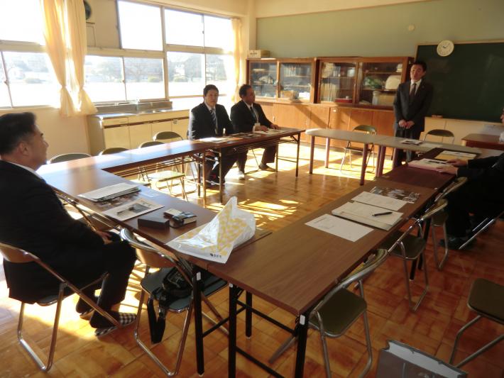 教室に4つの机が向い合せに並べられスーツを着た短髪の男性が手前に1人座っており、その奥に3人いる。うち1人は立ち2人は座っている