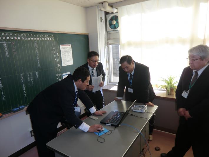 教室の黒板の前で、黒いスーツを着た短髪の男性が机を囲んで立っている。そのうち3人が机上のパソコンをのぞき込んでいる。