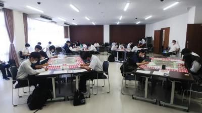 大会議室を4つの島に分け、赤と白のチェックのテーブルクロスをひいた机を並べ、生徒が4人から5人ずつ座り作業をしている写真