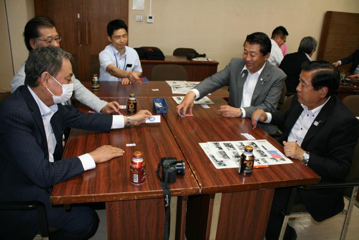 会議室に机が置かれ、スーツを着た短髪の男性5人が机を囲み座っている。机にはチラシが二枚置いてある