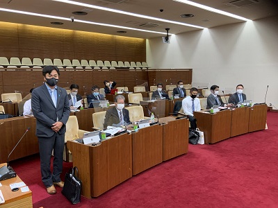 最前列の議席脇で起立してあいさつをする宜野湾市議会の男性委員長(石川委員長)。