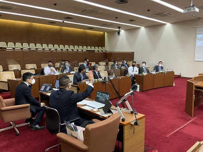 質疑席に座って議場スクリーンを指しながら説明する議会事務局職員。宜野湾市議会の皆さんは議席に座って聞いている。