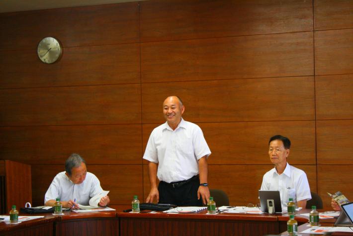 茶色い壁を背景に、白いワイシャツを着た男性が笑顔で立っている。その両脇に、白いワイシャツを着た短髪の男性が1人ずつ座っている