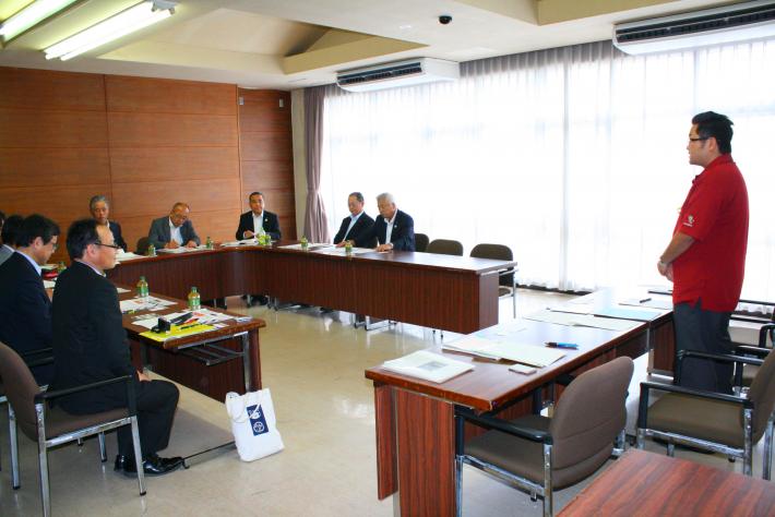 会議室内に、縦長の机がコの字型に置かれ、スーツを着た短髪の男性7名が座っている。手前に赤いポロシャツを着た男性が立っている
