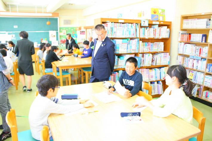 取手西小学校5年生図書室での授業を視察する議員。子どもたちがテーブルに座っている間を議員がぬうように歩いている。
