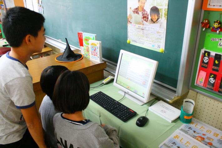 黒板の前に置かれたパソコンで、ほんくるのシステムを利用する小学生3人の写真
