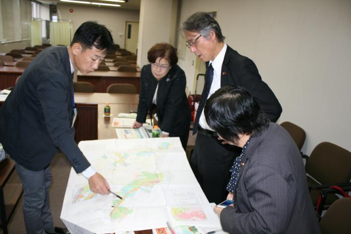 縦長の机に大きな紙の地図が置かれ、それをスーツを着た男性2人と女性2人が囲んで見ている。うち、男性1人がペンで地図を指している