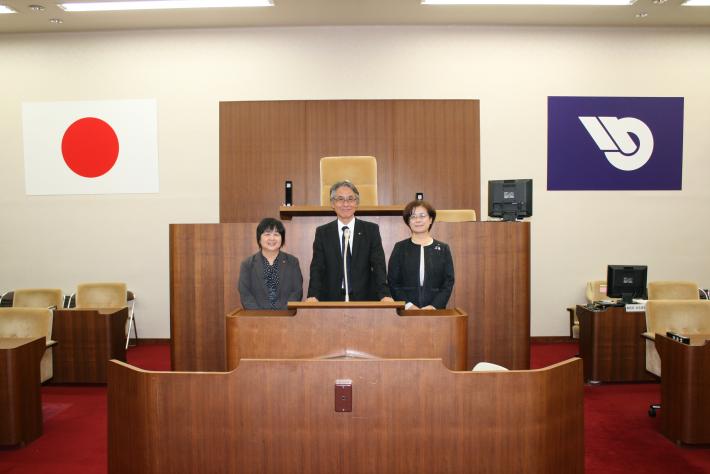 議場の議長席前にて、黒いスーツを着た男性1人と女性2人が横一列に並び、笑顔でこちらを見ている