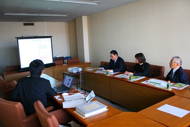 机がコの字型に置かれ、黒いスーツを着た男性3人と女性1人が座っている。奥にはスクリーンが設置されている