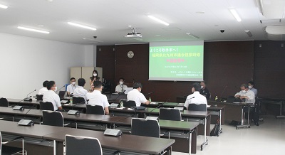 会議室内に6名のスーツを着た男性が背を向けて座り、奥にスーツの男性5名と女性1名がいる