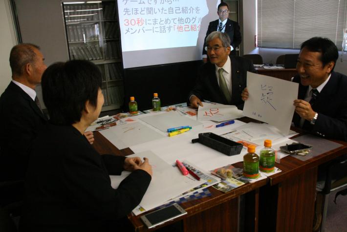 4人で1つのグループになって座っている。1人の男性議員が紙に書いた漢字1文字を他の3人に見せながら自己紹介をしている写真。