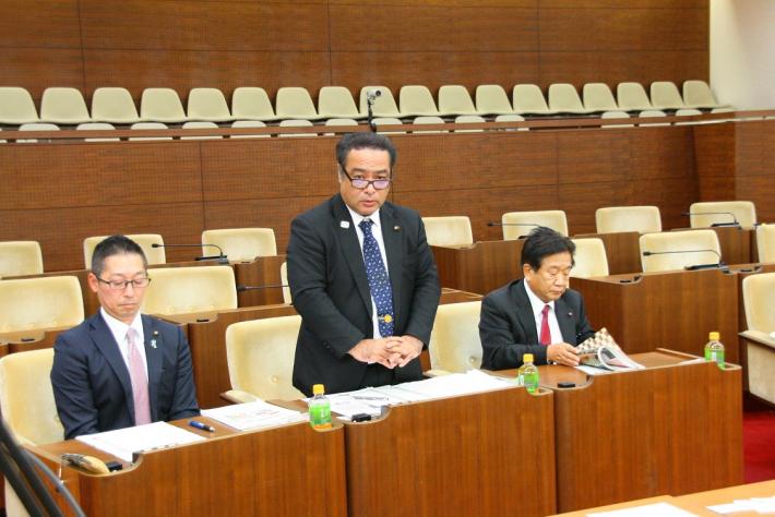 議席に3人の議員が座っている。真ん中の村上市男性議員(河村副委員長)が起立して視察のお礼を述べている写真