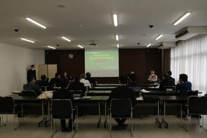 大会議室後方から撮った写真。スクリーンに緑色の資料が映る。参加者は横4列縦4列に並べれられた机椅子に座り正面を向いている