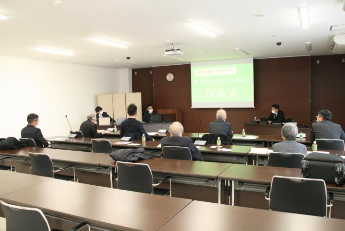 大会議室後方から撮った写真。スクリーンには緑色の資料が映る。会議室参加者は横4列縦2列に並べれられた机椅子に座り正面を向いている