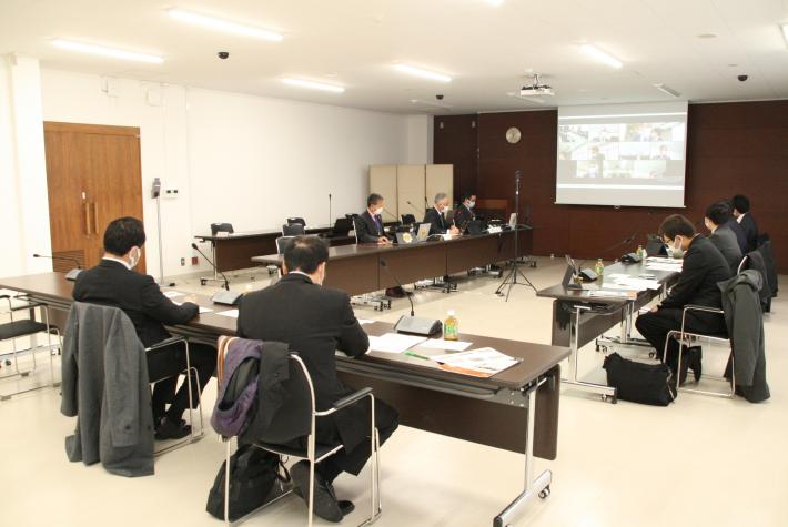 大会議室にて正面のスクリーンにオンライン会議の画面が映る。コの字型に並べられた長机には男性が8名座っている。