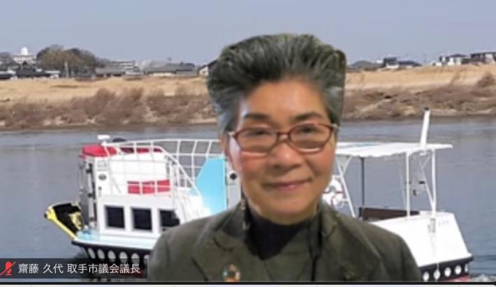Zoom画面に灰色の上着を着た短髪の女性が笑顔映っている。女性の背後には川を走る舟のバーチャル背景が映っている